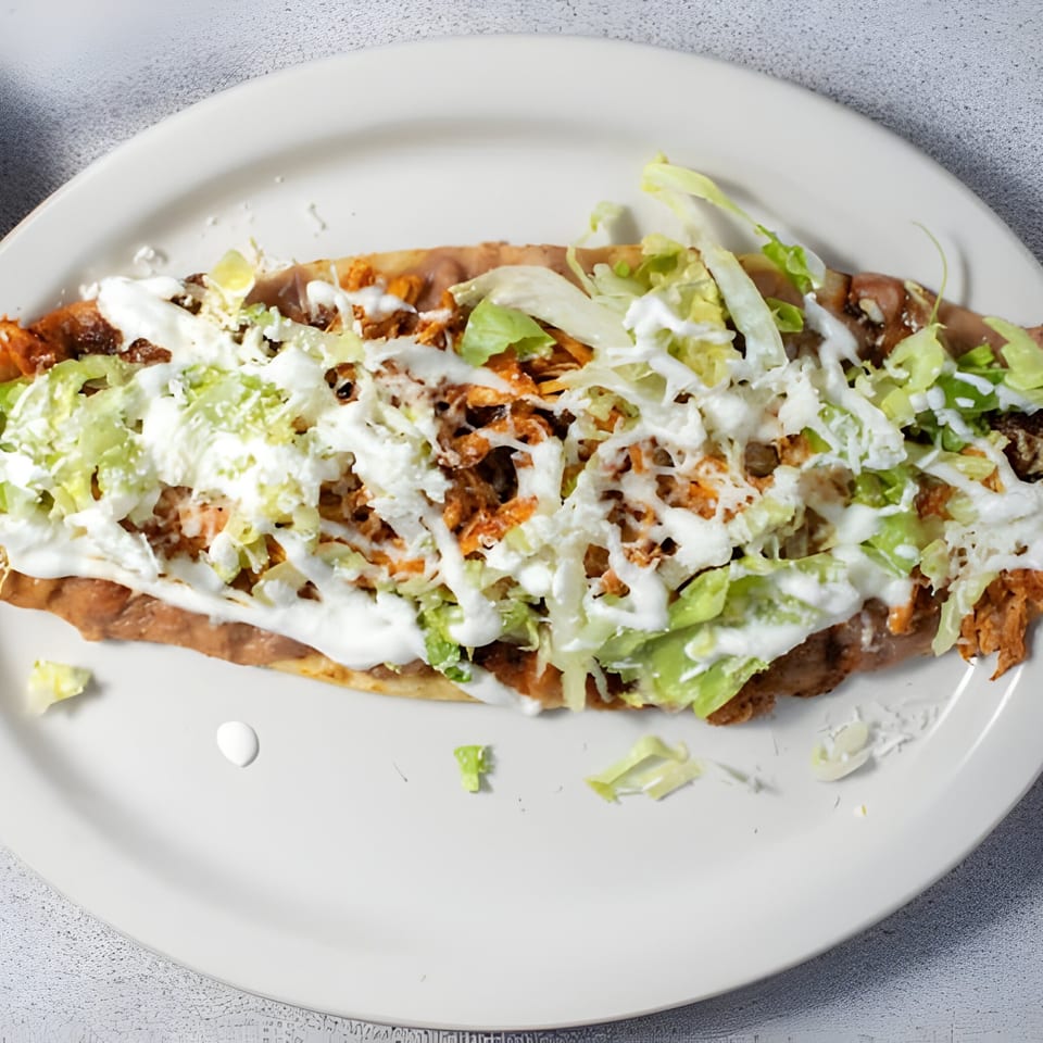 A Mexican huarache topped with refried beans, shredded meat, lettuce, sour cream, and cheese.