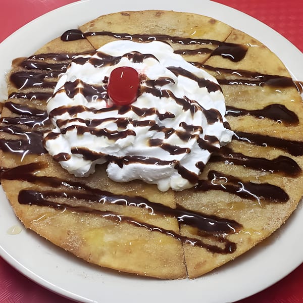A Mexican dessert with a flat fried pastry, whipped cream, chocolate drizzle, and a cherry.