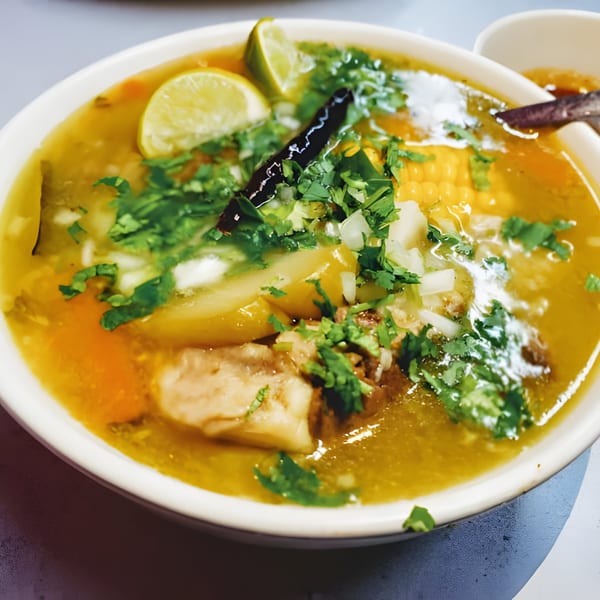 Close-up of a Mexican soup with meat, corn, carrots, cilantro, and lime wedges.