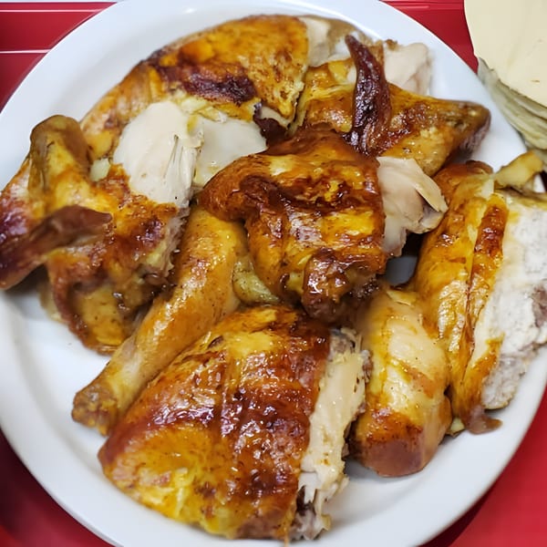 A white plate filled with pieces of golden-brown rotisserie chicken, with tortillas in the background.